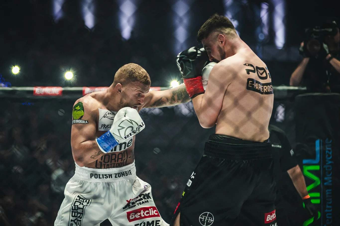 Dwóch zawodników MMA walczy w oktagonie, obaj skupieni na wymianie ciosów, tłum widzów obecny w tle, wyraźne oznaczenia sponsorów na spodenkach i bandach wokół ringu.