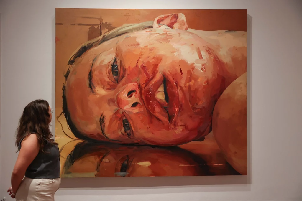 Wystawa obrazów Jenny Saville w Londynie Duży obraz malarski prezentujący zdeformowaną i poranioną twarz człowieka z widocznymi śladami krwi, odbitą w błyszczącej powierzchni, obok obrazu stoi kobieta oglądająca dzieło.