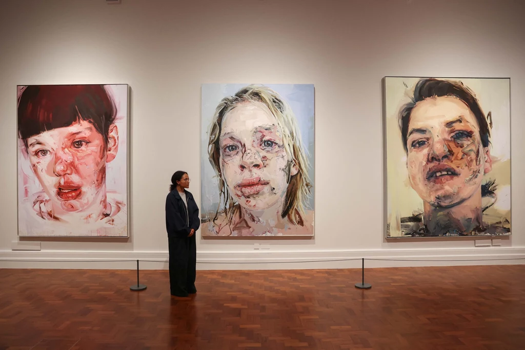 Wystawa The anatomy of painting Jenny Saville Przestronne wnętrze galerii sztuki z trzema dużymi, ekspresyjnymi portretami współczesnymi na ścianie; kobieta w ciemnym ubraniu stoi przed obrazami, analizując ich szczegóły, podłoga z drewna, jasna kolorystyka ścian.