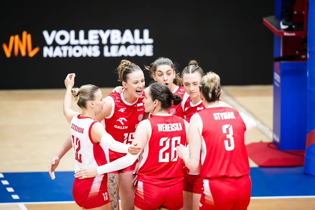 Grupa siatkarek w czerwonych strojach drużyny narodowej Polski świętuje punkt podczas meczu siatkarskiej Ligi Narodów, wokół widoczne elementy boiska oraz logo turnieju na czarnym tle.