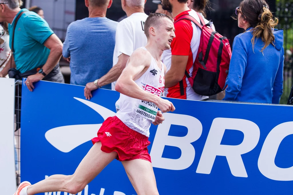 Marcin Chabowski Biegacz w stroju sportowym i z numerem startowym pokonuje trasę zawodów, w tle widzowie przy barierce kibicują uczestnikom maratonu.