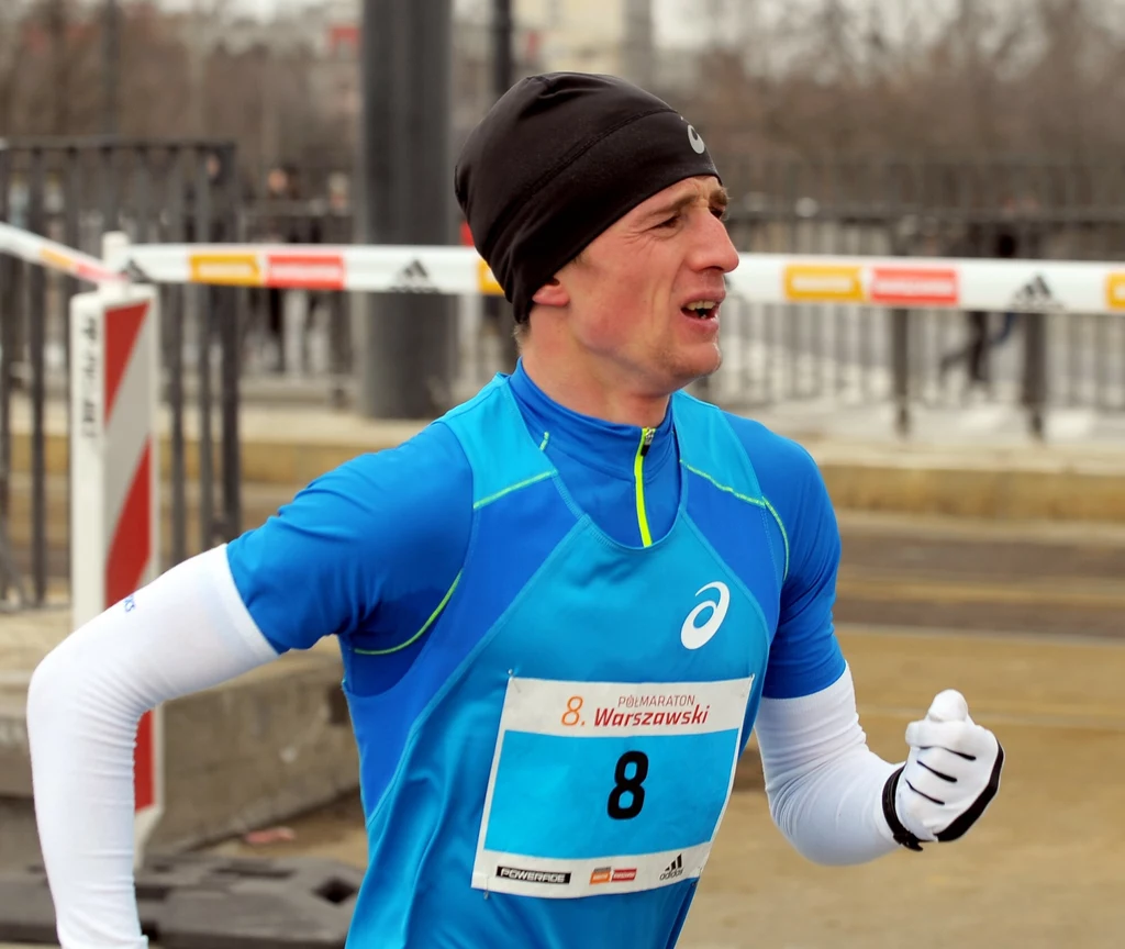 Marcin Chabowski Mężczyzna w sportowym stroju biegnie ulicą podczas zawodów biegowych z numerem startowym 8, za nim widoczne są bariery zabezpieczające trasę.