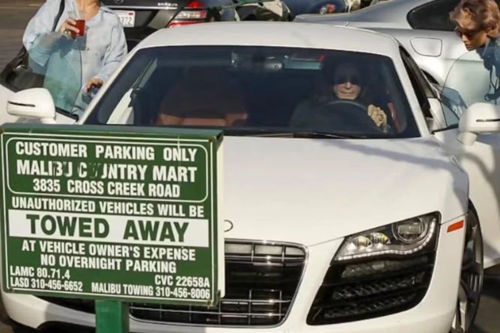 Biały samochód marki Audi zaparkowany na parkingu przed tablicą informacyjną o zasadach parkowania, w samochodzie siedzi kobieta w okularach przeciwsłonecznych, za samochodem widoczne inne pojazdy oraz osoby na parkingu.
