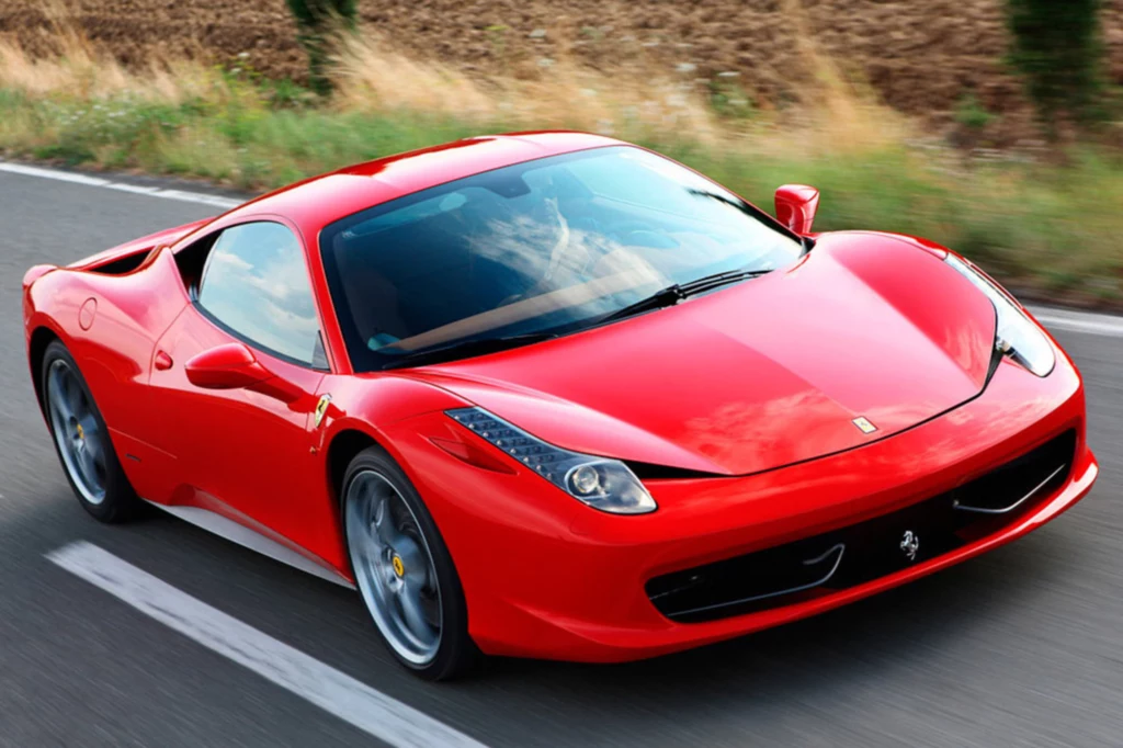Czerwone Ferrari jedzie po pustej, asfaltowej drodze, otoczonej przez pole i zieleń, pojazd prezentuje sportowy, aerodynamiczny kształt i błyszczący lakier.