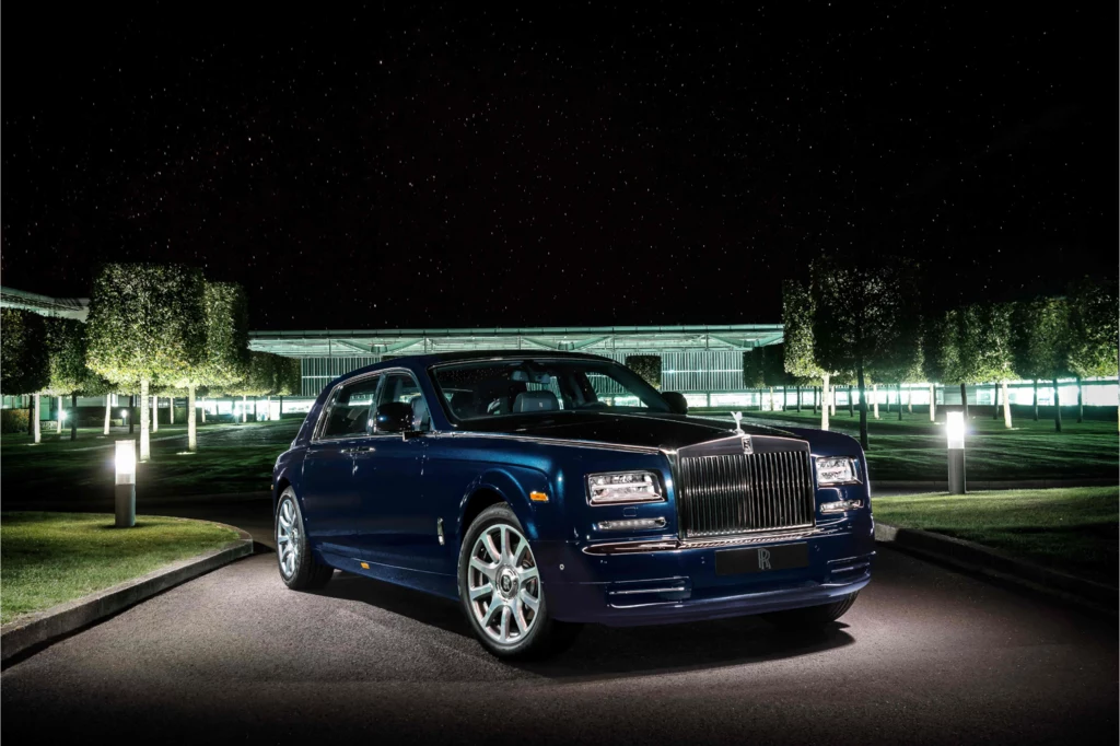 Luksusowy, ciemnogranatowy samochód marki Rolls-Royce zaparkowany nocą na podjeździe, otoczony eleganckimi, przystrzyżonymi drzewami i podświetlonymi alejkami, w tle nowoczesny budynek o szklanej fasadzie.