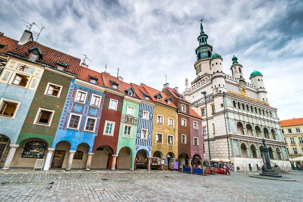 Kolorowe kamieniczki z arkadami oraz okazały renesansowy ratusz z wysoką wieżą, otaczające brukowany rynek pod zachmurzonym niebem.