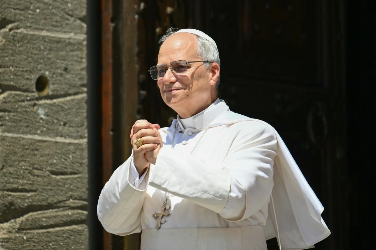 Papież Leon XIV ogłosił cud w amerykańskim szpitalu Mężczyzna w białym stroju papieskim z krzyżem na szyi stoi na tle kamiennych drzwi, dłonie trzyma złożone w geście modlitwy lub skupienia, delikatnie uśmiechnięty, z okularami i pierścieniem na palcu.