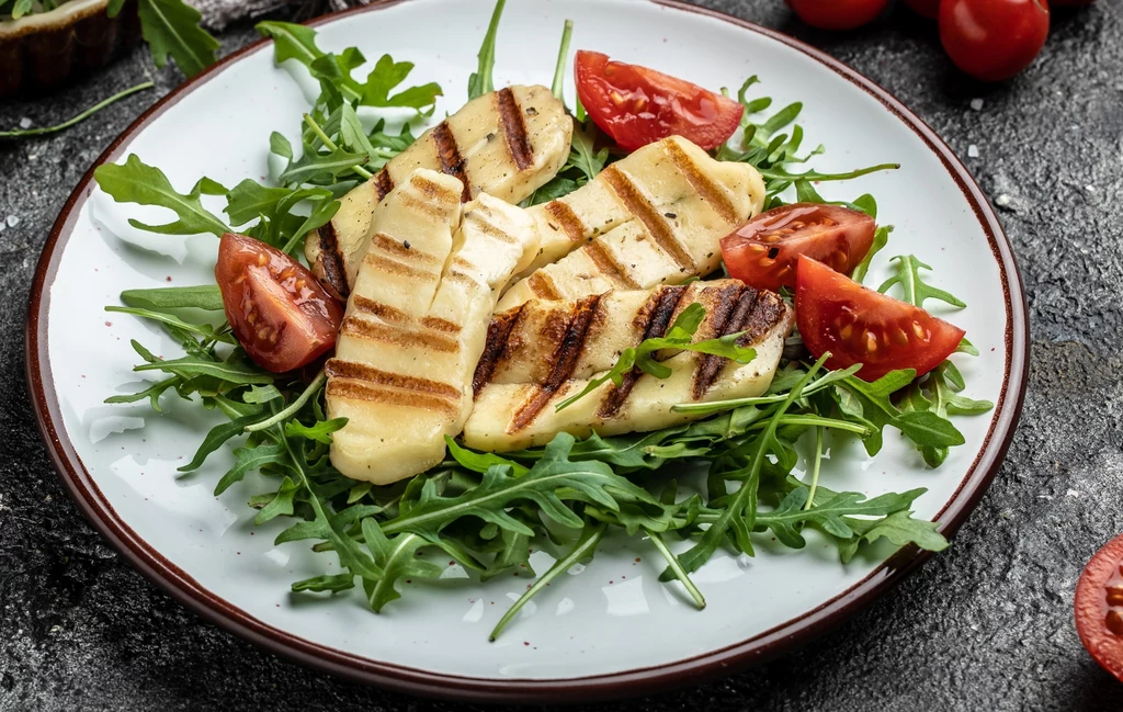 Grillowany ser halloumi ułożony na świeżej rukoli, dopełniony cząstkami pomidorów, całość podana na białym talerzu na ciemnym tle.
