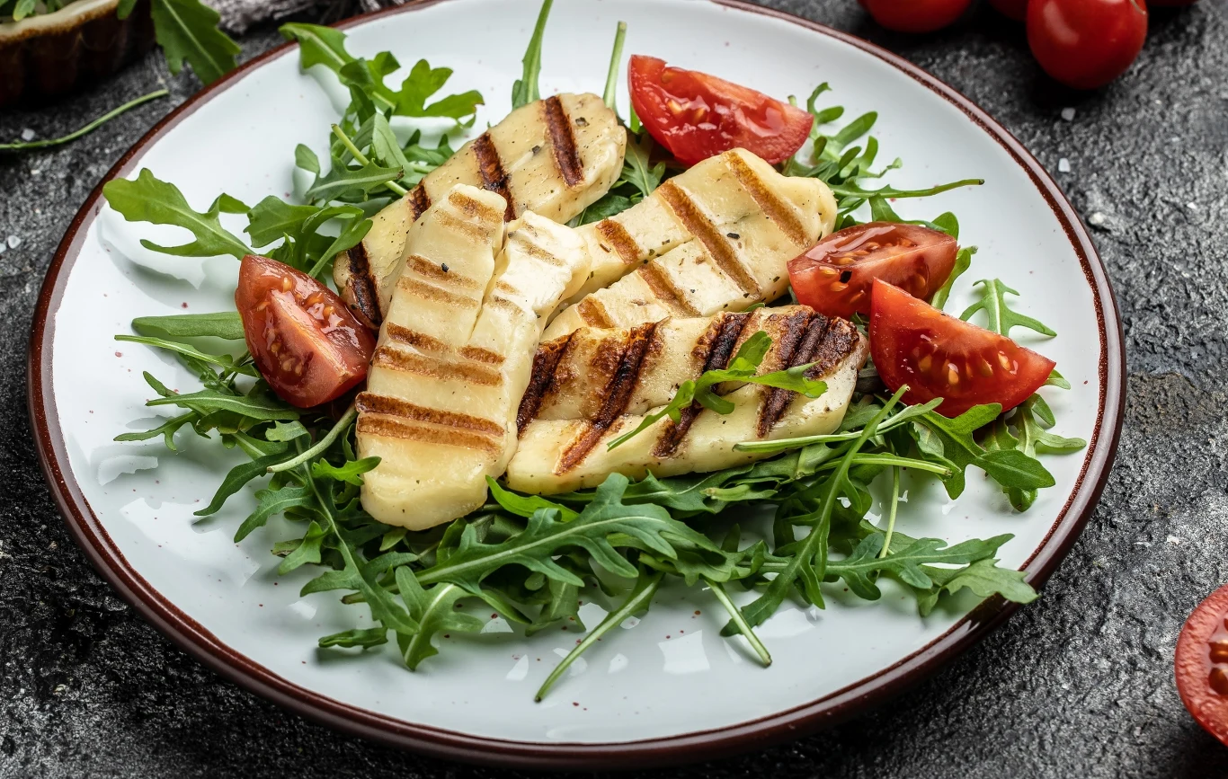 Grillowany ser halloumi ułożony na świeżej rukoli, dopełniony cząstkami pomidorów, całość podana na białym talerzu na ciemnym tle.