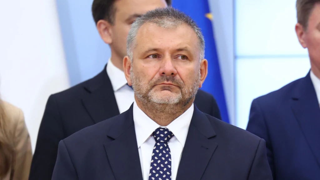 Waldemar Żurek został nowym ministrem sprawiedliwości Mężczyzna w formalnym garniturze i krawacie z widoczną siwizną i krótką brodą stoi na tle innych osób w podobnych strojach, w tle widoczny fragment flagi i biało-niebieskie wnętrze.