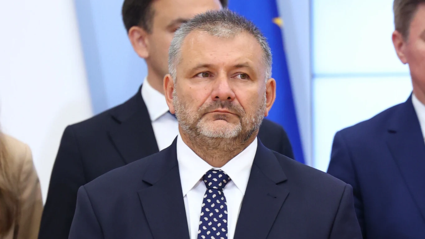 Mężczyzna w formalnym garniturze i krawacie z widoczną siwizną i krótką brodą stoi na tle innych osób w podobnych strojach, w tle widoczny fragment flagi i biało-niebieskie wnętrze.