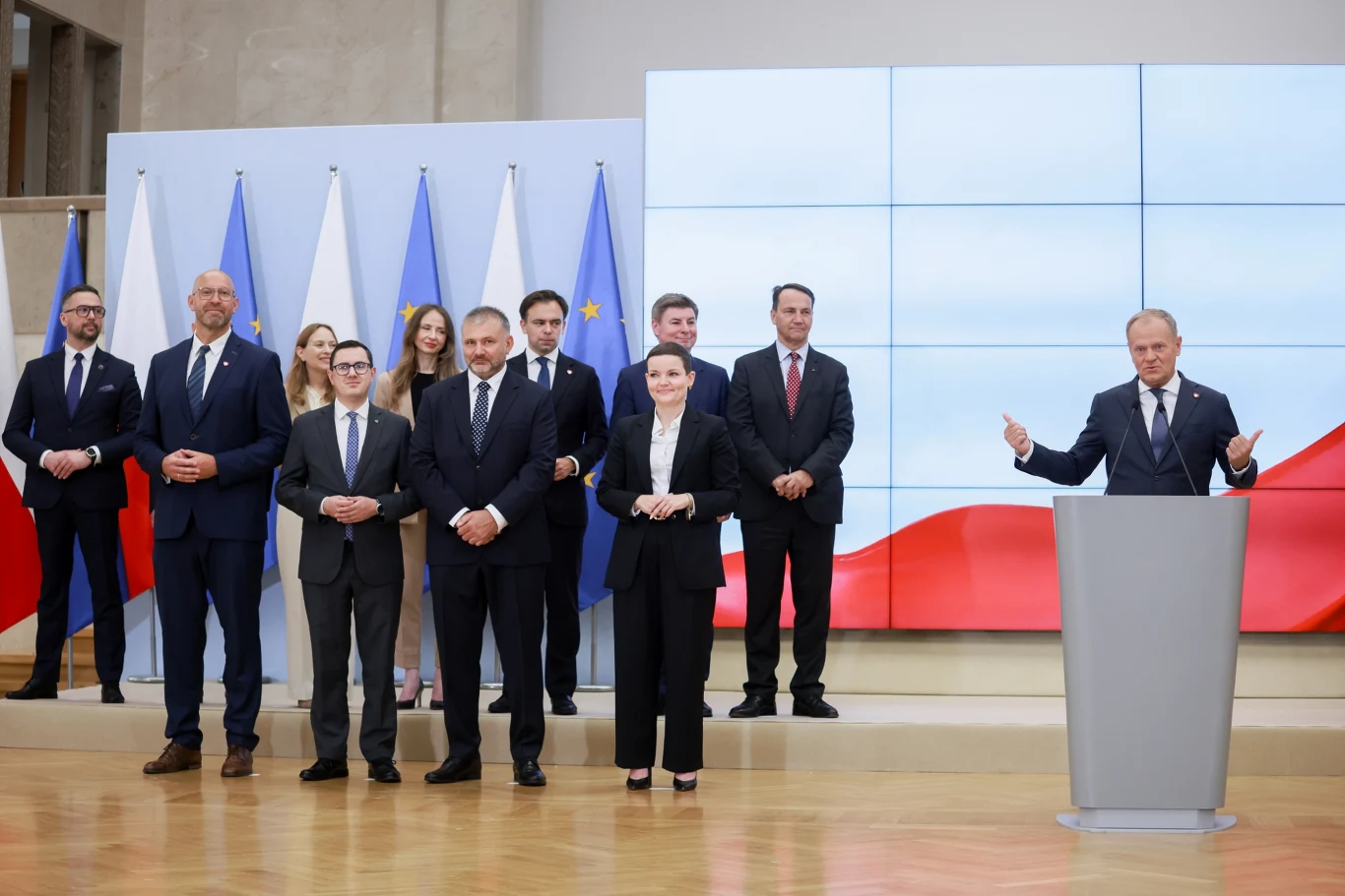 Donald Tusk ogłasza szczegóły rekonstrukcji rządu, w pierwszym rzędzie od lewej nowi ministrowie: sportu - Jakub Rutnicki, energii Miłosz Motyka, sprawiedliwości - Waldemar Żurek, kultury - Marta Cienkowska Grupa elegancko ubranych osób stojących na tle flag Polski i Unii Europejskiej, mężczyzna przemawiający przy mównicy, w tle duży ekran z polską flagą, atmosfera oficjalna i profesjonalna.