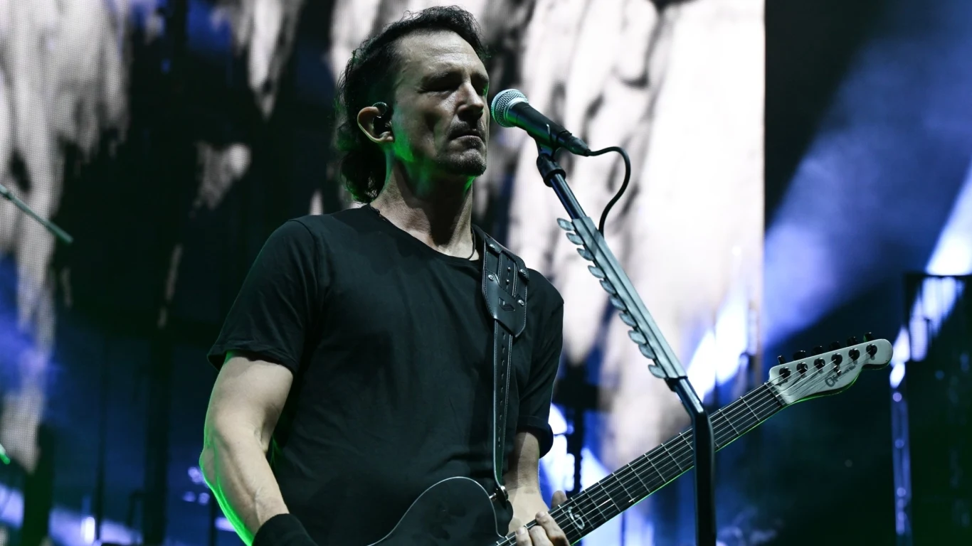 Joe Duplantier (Gojira) Muzyk ubrany na czarno grający na gitarze elektrycznej i śpiewający do mikrofonu na tle sceny oświetlonej kolorowymi światłami i abstrakcyjną grafiką.