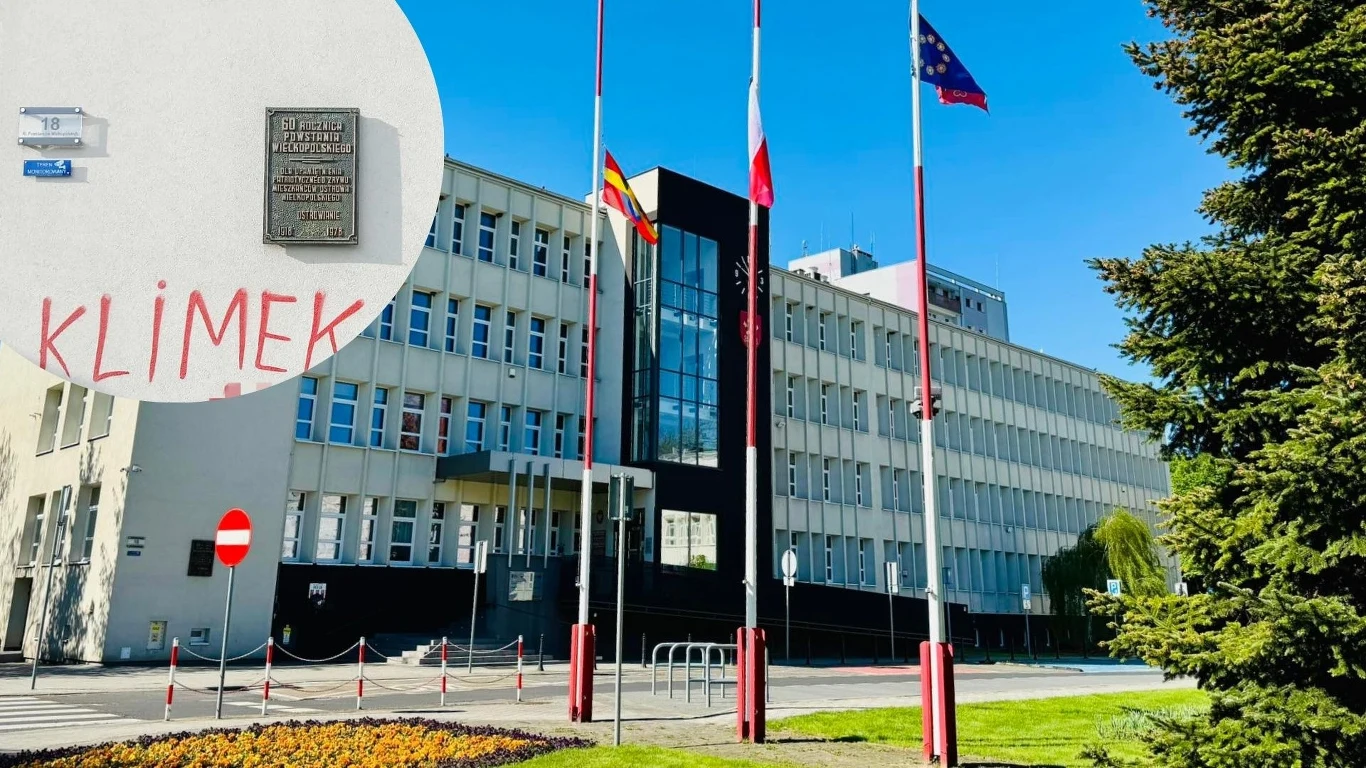 Akt wandalizmu w Ostrowie Wielkopolskim. Policja szuka sprawców (zdj. ilustracyjne) Nowoczesny, urzędowy budynek z flagami Polski, Unii Europejskiej i regionu, widoczne uporządkowane otoczenie z kwiatami, w lewym górnym rogu zbliżenie na ścianę z tablicą pamiątkową i napisem sprayem Klimek.