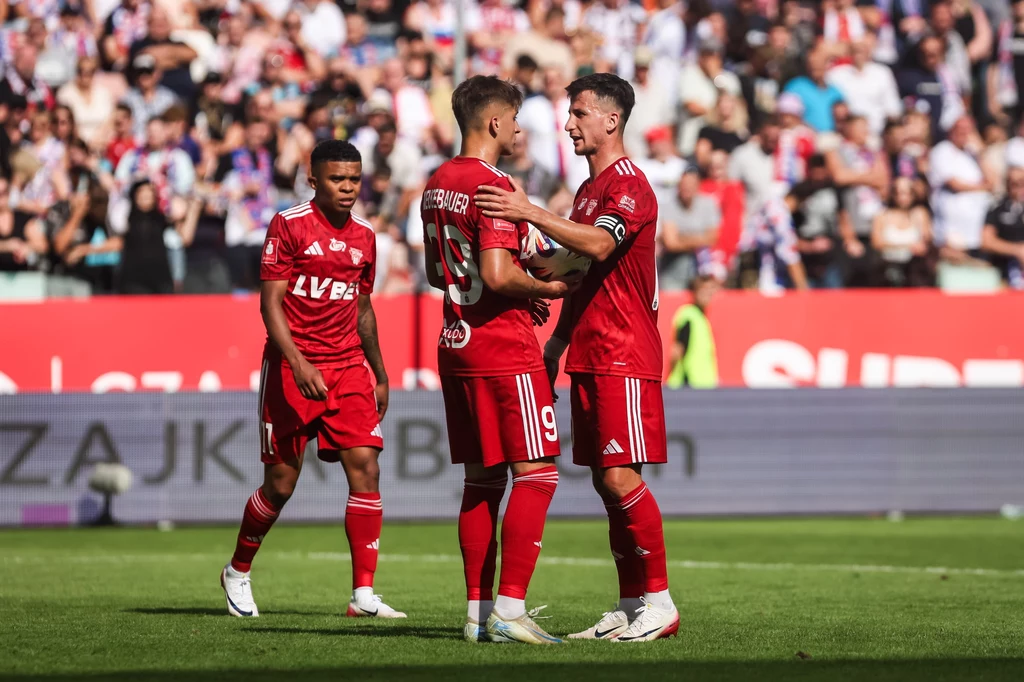 Piłkarze Lechii Gdańsk w meczu Ekstraklasy z Górnikiem Zabrze Trzech piłkarzy w czerwonych strojach drużyny LV BET stoi na boisku piłkarskim, dwóch z nich wymienia gesty wsparcia, wokół widzowie na trybunach tworzą tło wydarzenia sportowego.