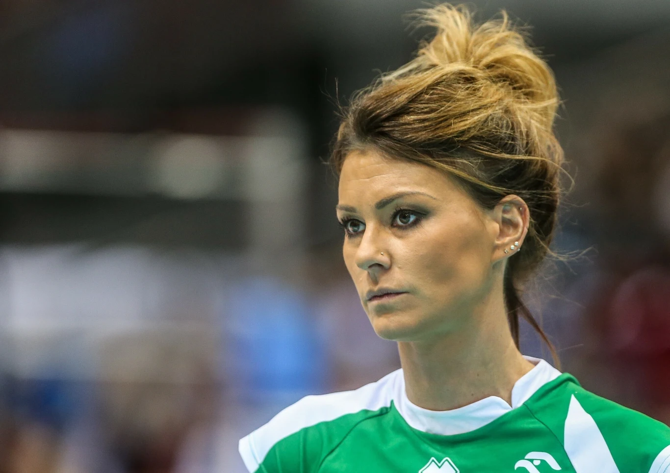 Kobieta w sportowej zielonej koszulce z logo, z upiętymi włosami, patrząca poważnie w bok, w tle rozmyte postacie i hala sportowa.