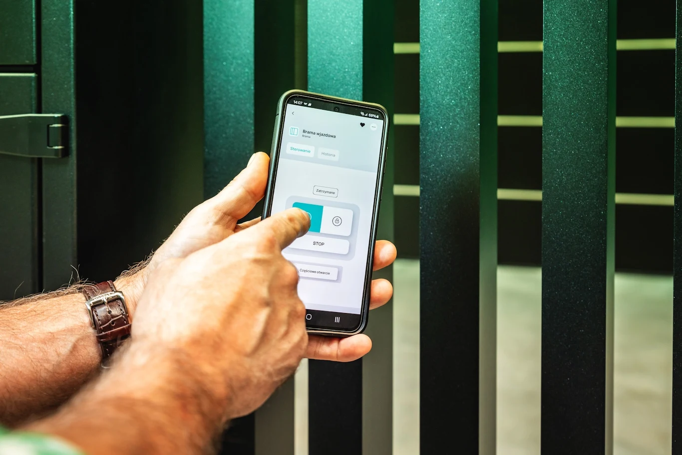 Mężczyzna obsługuje bramę lub drzwi za pomocą aplikacji na smartfonie, naciskając przycisk na ekranie, w tle widoczne metalowe ogrodzenie.
