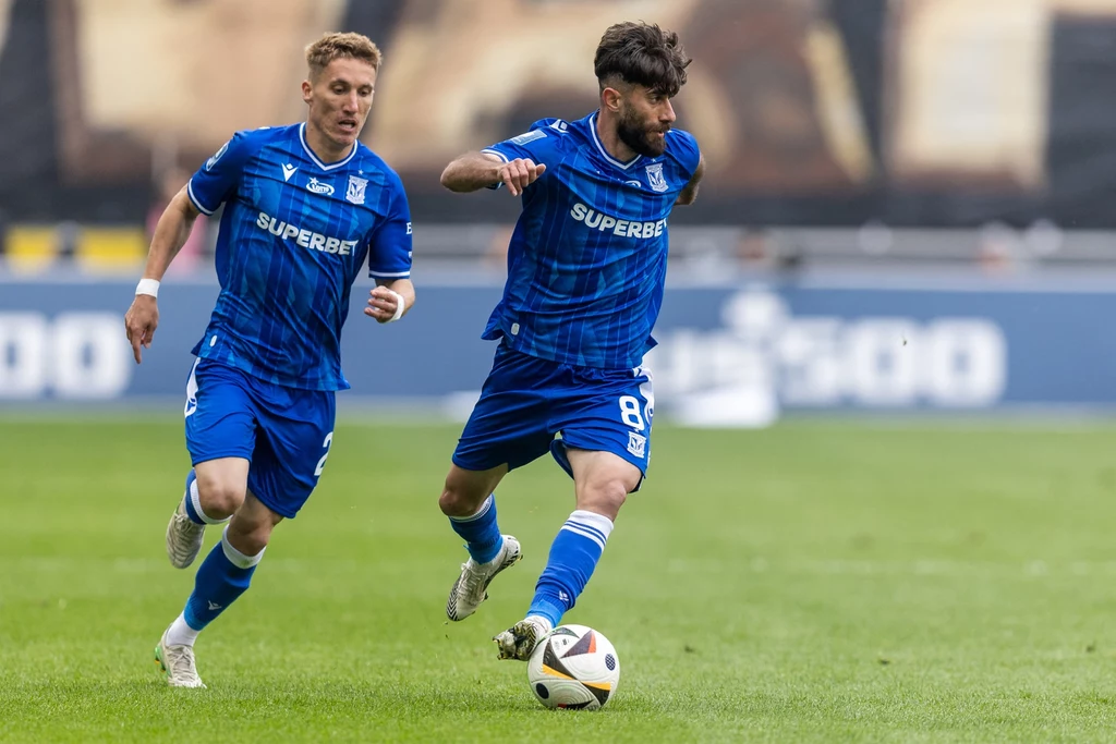Joel Pereira i Ali Gholizadeh w barwach Lecha Poznań Dwóch piłkarzy w niebieskich strojach drużyny Lech Poznań biegnących po murawie podczas meczu, jeden z nich prowadzi piłkę przy nodze, drugi podąża tuż obok niego.