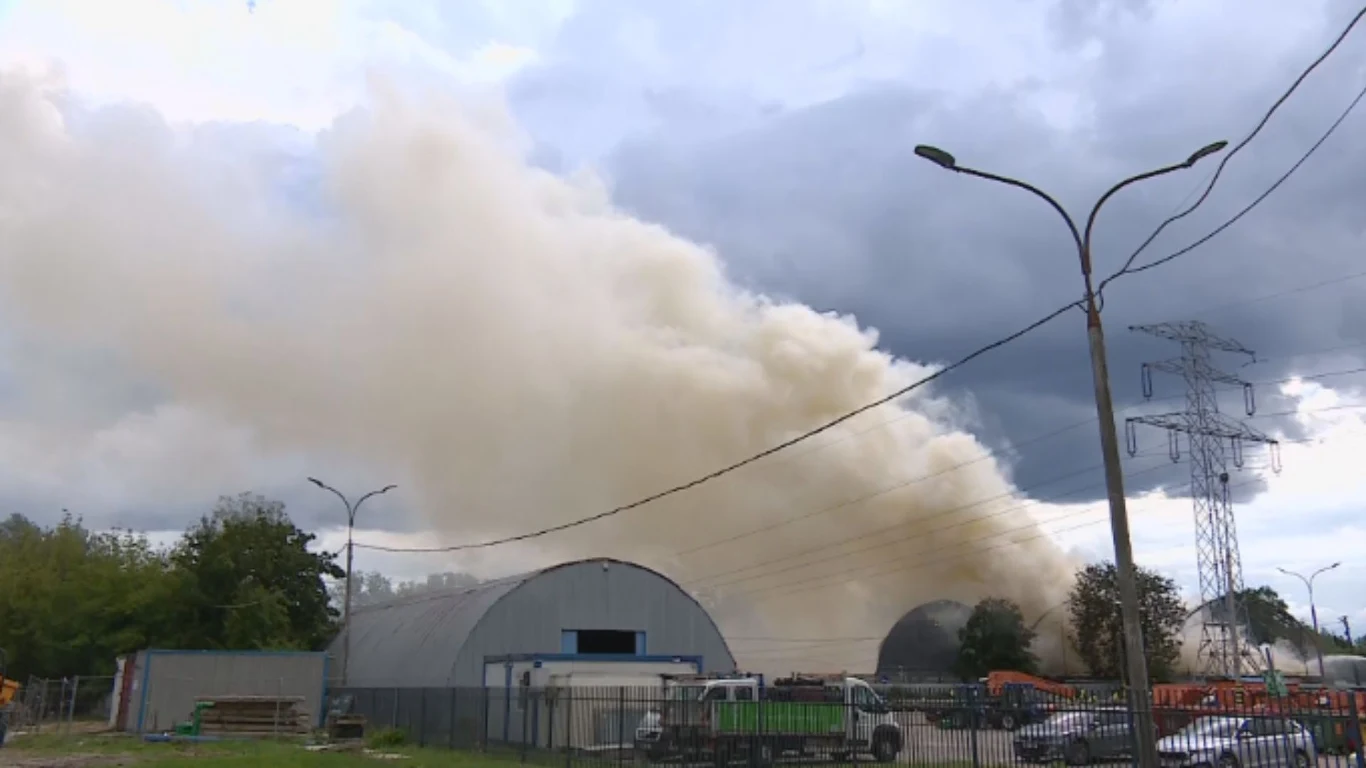 Gęste kłęby białego dymu unoszące się nad dużym magazynem otoczonym przez pojazdy i ogrodzenie, w tle pochmurne niebo i linie wysokiego napięcia dodające dramatyzmu scenie.