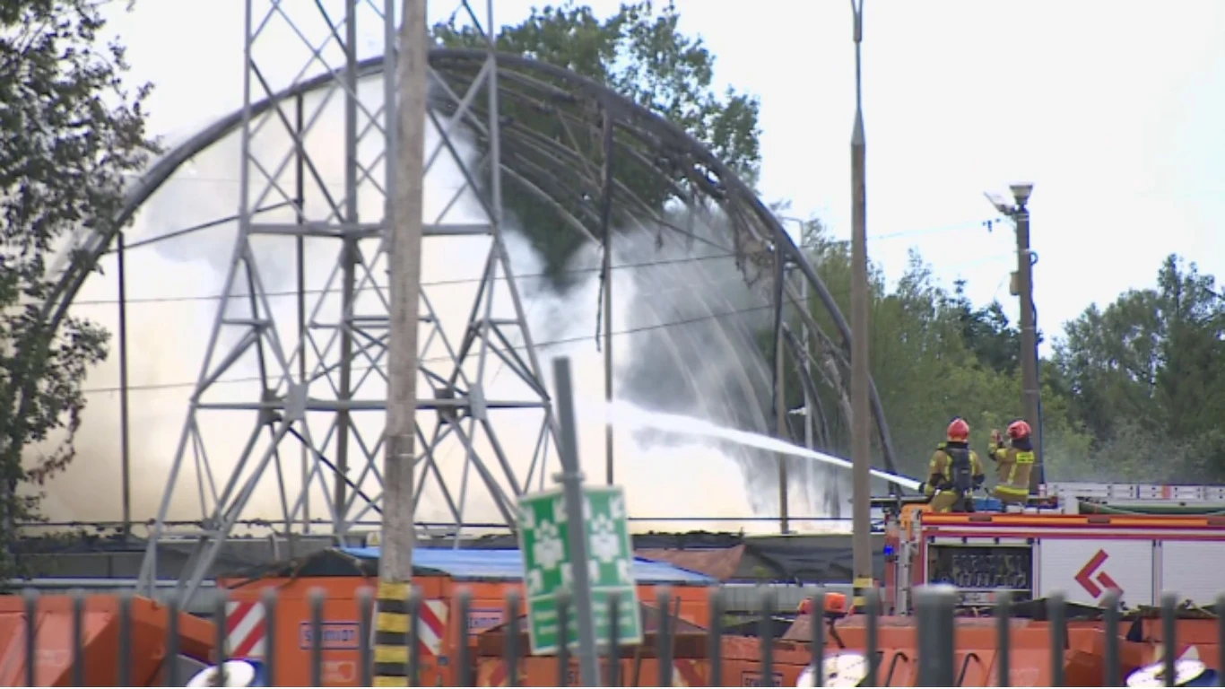 Strażacy gaszą pożar w dużym obiekcie przemysłowym, wokół znajdują się kontenery i sprzęt budowlany, w tle widoczny dym i konstrukcja metalowa.