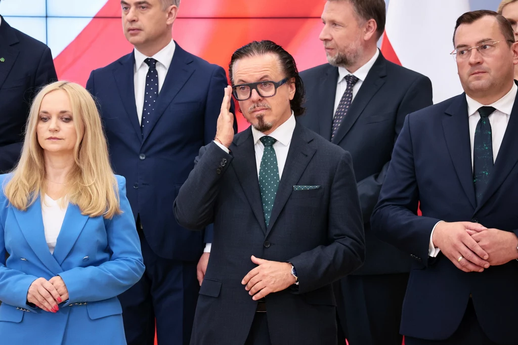 Wojciech Balczun ma bardzo duże doświadczenie na kierowniczych stanowiskach w państwowych spółkach Grupa elegancko ubranych osób stojących razem, większość w ciemnych garniturach, jedna kobieta w niebieskim garniturze, centralna postać w okularach dotyka twarzy, w tle widoczna biało-czerwona flaga.