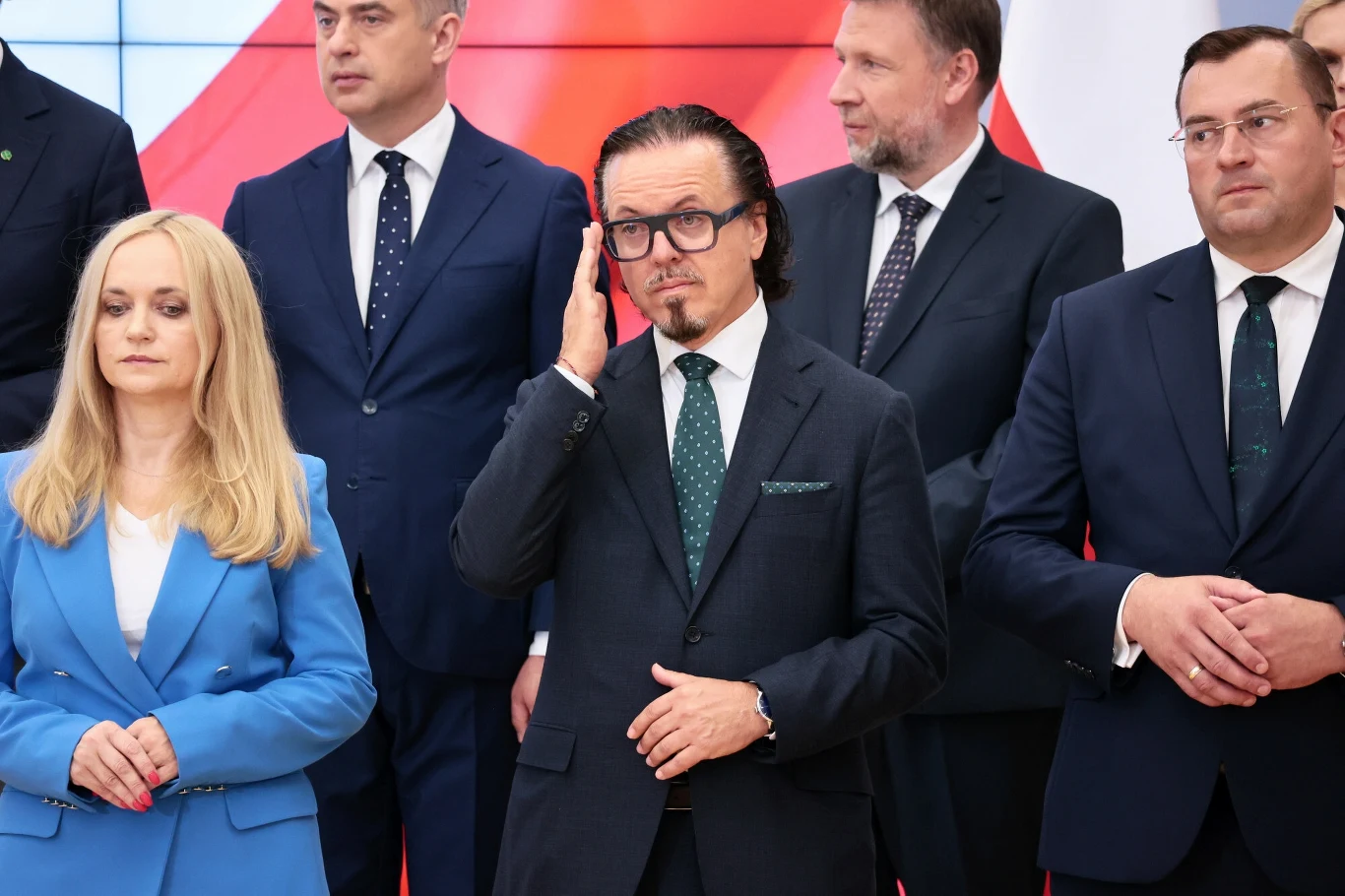 Grupa elegancko ubranych osób stojących razem, większość w ciemnych garniturach, jedna kobieta w niebieskim garniturze, centralna postać w okularach dotyka twarzy, w tle widoczna biało-czerwona flaga.