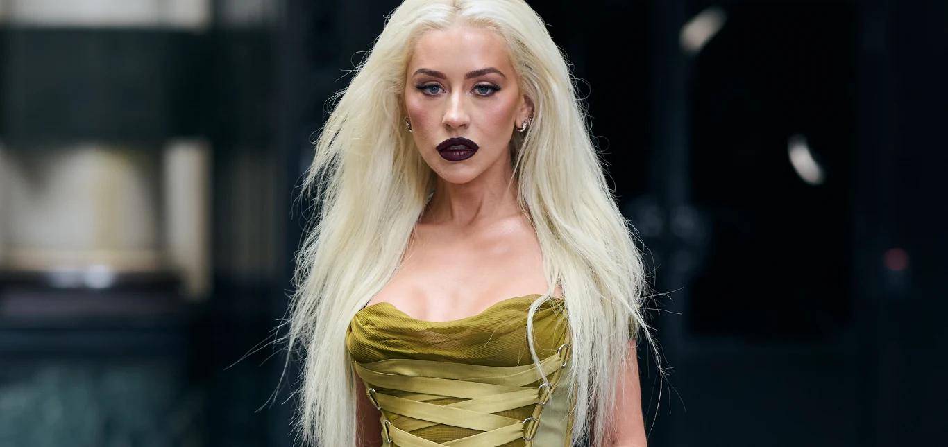 Christina Aguilera Kobieta z długimi, platynowymi blond włosami i ciemną szminką na ustach ma na sobie oliwkowozieloną suknię z gorsetowym topem, stoi w miejskim otoczeniu.