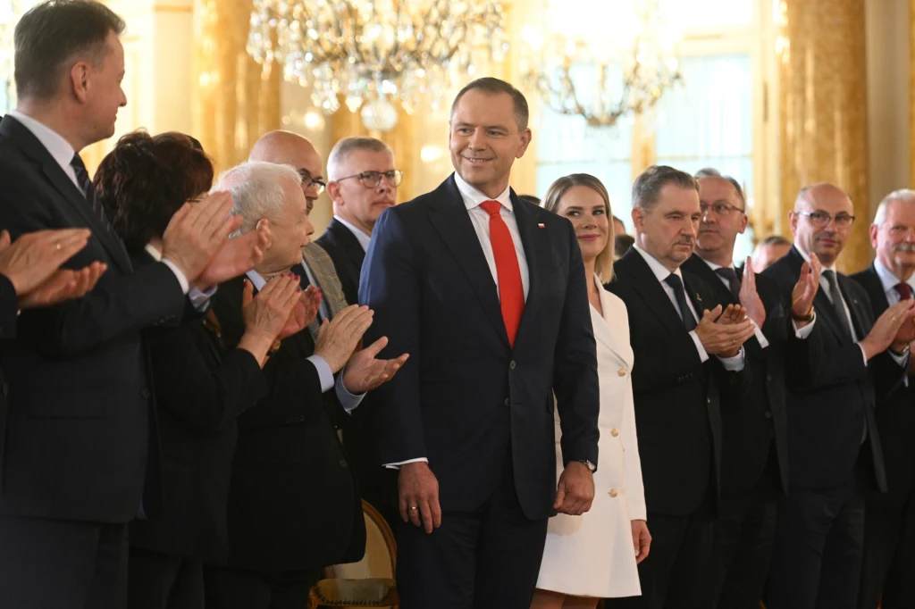 Wręczenie zaświadczenia o wyborze na Prezydenta RP Karolowi Nawrockiemu Grupa elegancko ubranych osób uczestniczy w oficjalnym wydarzeniu w reprezentacyjnej sali z żyrandolami, centralną postacią jest mężczyzna w granatowym garniturze i czerwonym krawacie otoczony przez gratulujących mu ludzi.