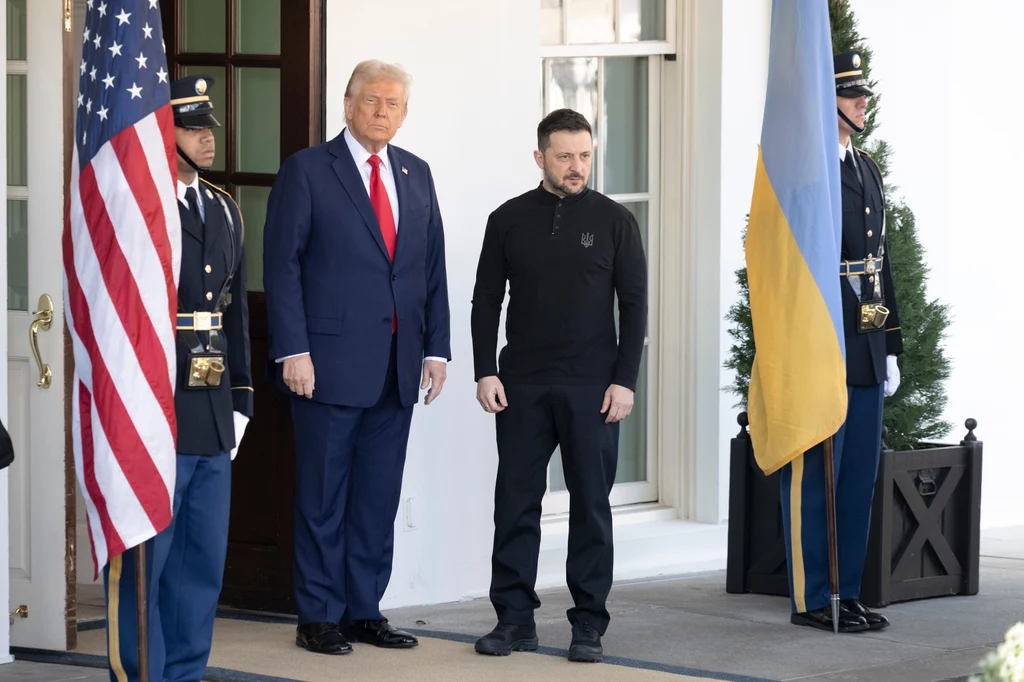 W lutym prezydent USA Donald Trump spotkał się z prezydentem Ukrainy Wołodymyrem Zełenskim w Białym Domu Dwóch mężczyzn stoi obok siebie na tle flag Stanów Zjednoczonych i Ukrainy, po bokach stoją strażnicy w mundurach. Mężczyzna po lewej ubrany jest w garnitur, a po prawej w ciemny strój.