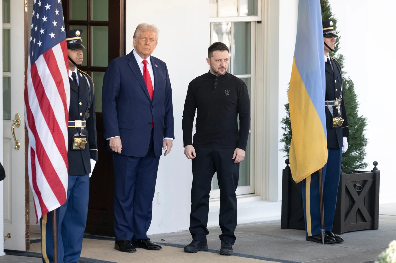 Dwóch mężczyzn stoi obok siebie na tle flag Stanów Zjednoczonych i Ukrainy, po bokach stoją strażnicy w mundurach. Mężczyzna po lewej ubrany jest w garnitur, a po prawej w ciemny strój.