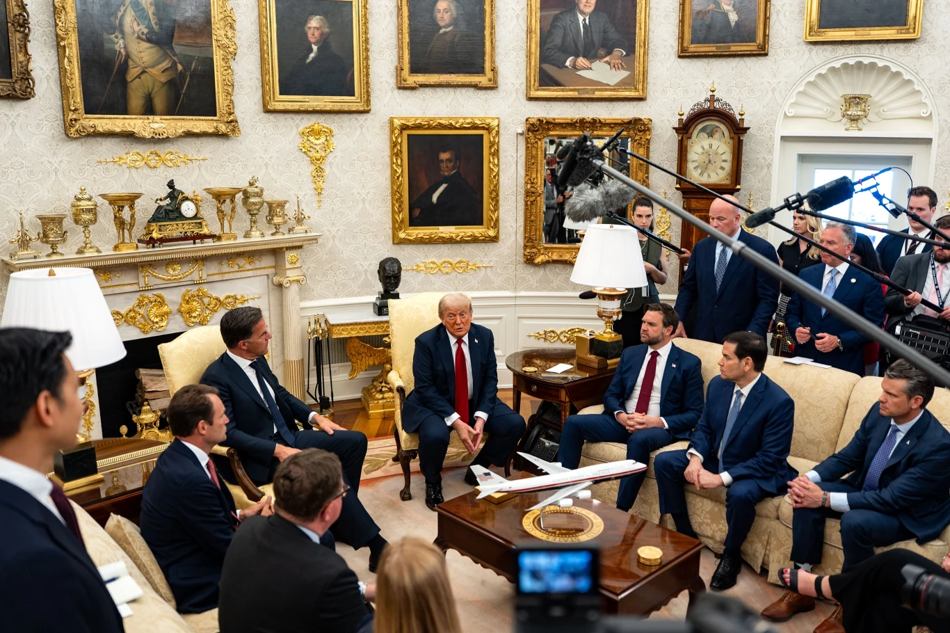 Oficjalne spotkanie polityczne w eleganckim gabinecie, gdzie centralną postacią jest mężczyzna w czerwonym krawacie otoczony przez grupę innych mężczyzn w garniturach, siedzących wokół stołu z modelem samolotu, otoczenie wypełnione portretami historycz...