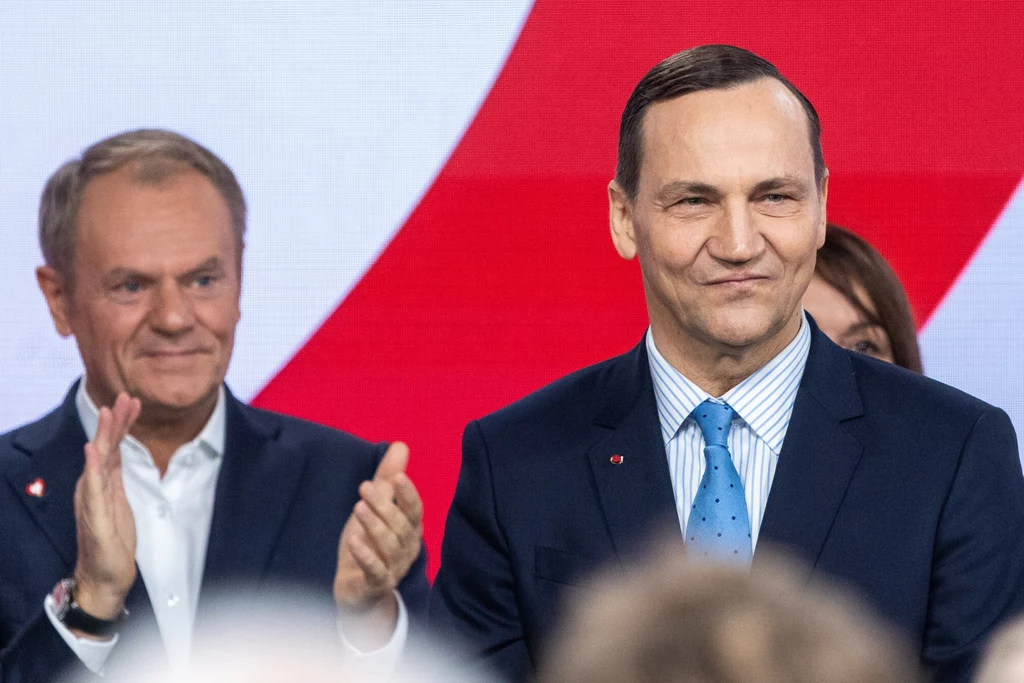 Dwóch mężczyzn w garniturach na tle flagi w biało-czerwonych barwach, jeden z nich stoi z przodu z lekkim uśmiechem, drugi bije brawo w tle, wyraz twarzy obu wyraża zadowolenie