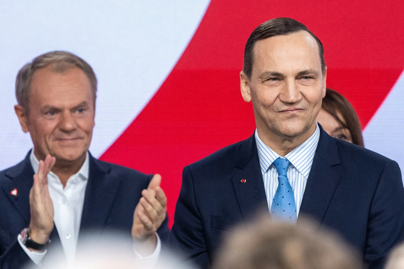 Donald Tusk odda swoje stanowisko Sikorskiemu. Oto powody Dwóch mężczyzn w garniturach na tle flagi w biało-czerwonych barwach, jeden z nich stoi z przodu z lekkim uśmiechem, drugi bije brawo w tle, wyraz twarzy obu wyraża zadowolenie