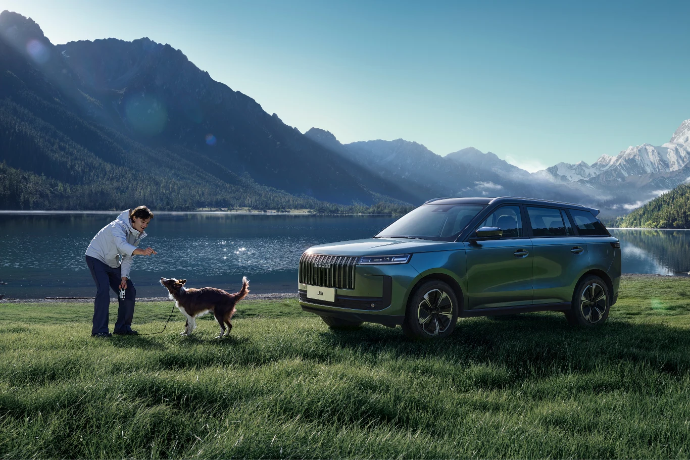 Mężczyzna bawiący się z psem na zielonej łące nad jeziorem w otoczeniu gór oraz zaparkowany nowoczesny samochód terenowy na pierwszym planie.