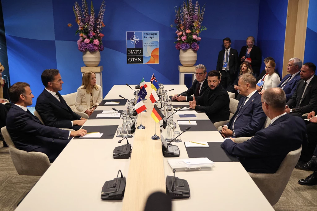 Zełenski i europejscy przywódcy podczas szczytu NATO w Hadze Grupa światowych przywódców siedzi wokół dużego stołu konferencyjnego podczas szczytu NATO, na stole flagi kilku państw europejskich, w tle logotyp NATO oraz dekoracje kwiatowe.