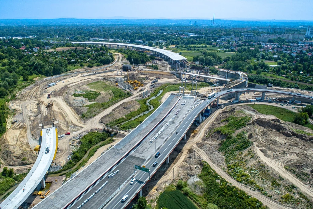 Trwa budowa 5 km drogi S7 od węzła Mistrzejowice do węzła Nowa Huta. Widok z góry na rozbudowaną autostradę oraz budowę nowych odcinków drogowych, liczne maszyny budowlane rozmieszczone wokół, zielone tereny w tle i zabudowania miejskie na horyzoncie.