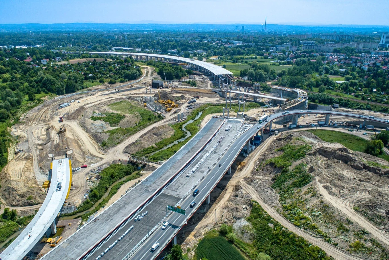Widok z góry na rozbudowaną autostradę oraz budowę nowych odcinków drogowych, liczne maszyny budowlane rozmieszczone wokół, zielone tereny w tle i zabudowania miejskie na horyzoncie.