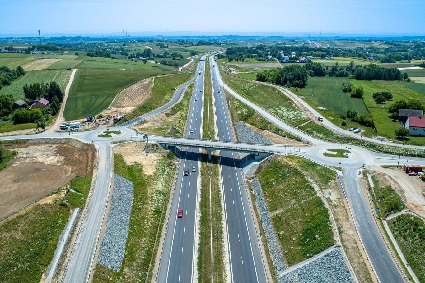 Autostrada przebiegająca przez tereny wiejskie, nad którą wybudowany jest most, po bokach rozciągają się pola uprawne, widoczne są pojedyncze domy oraz kilka samochodów poruszających się po jezdni.