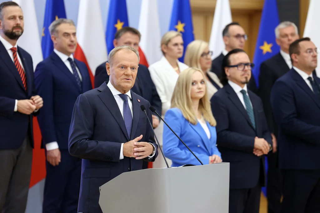 Premier Donald Tusk ogłasza zmiany w rządzie Grupa polityków ubrana w garnitury i eleganckie stroje stoi na tle flag Polski i Unii Europejskiej, mężczyzna na pierwszym planie przemawia do mikrofonu, obok niego kobieta w niebieskiej marynarce, pozostali uczestnicy stoją w rzędach.