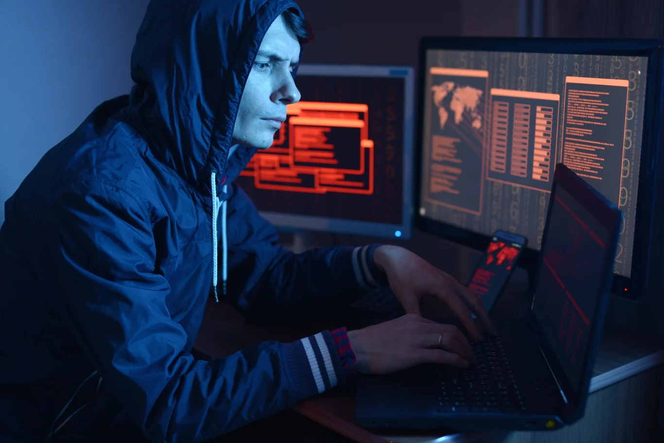 Ataki ransomware to prawdziwe wyzwanie dla systemów bezpieczeństwa Młody mężczyzna w bluzie z kapturem siedzący przy laptopie otoczonym monitorami z wyświetlonymi kodami i mapami, skupiony na pracy w przyciemnionym pomieszczeniu