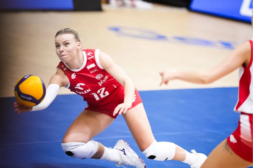 Siatkarka w czerwonym stroju z numerem 12 wykonuje dynamiczny ruch podczas próby przyjęcia piłki, skupiona na grze, w tle fragment innej zawodniczki i boisko sportowe.