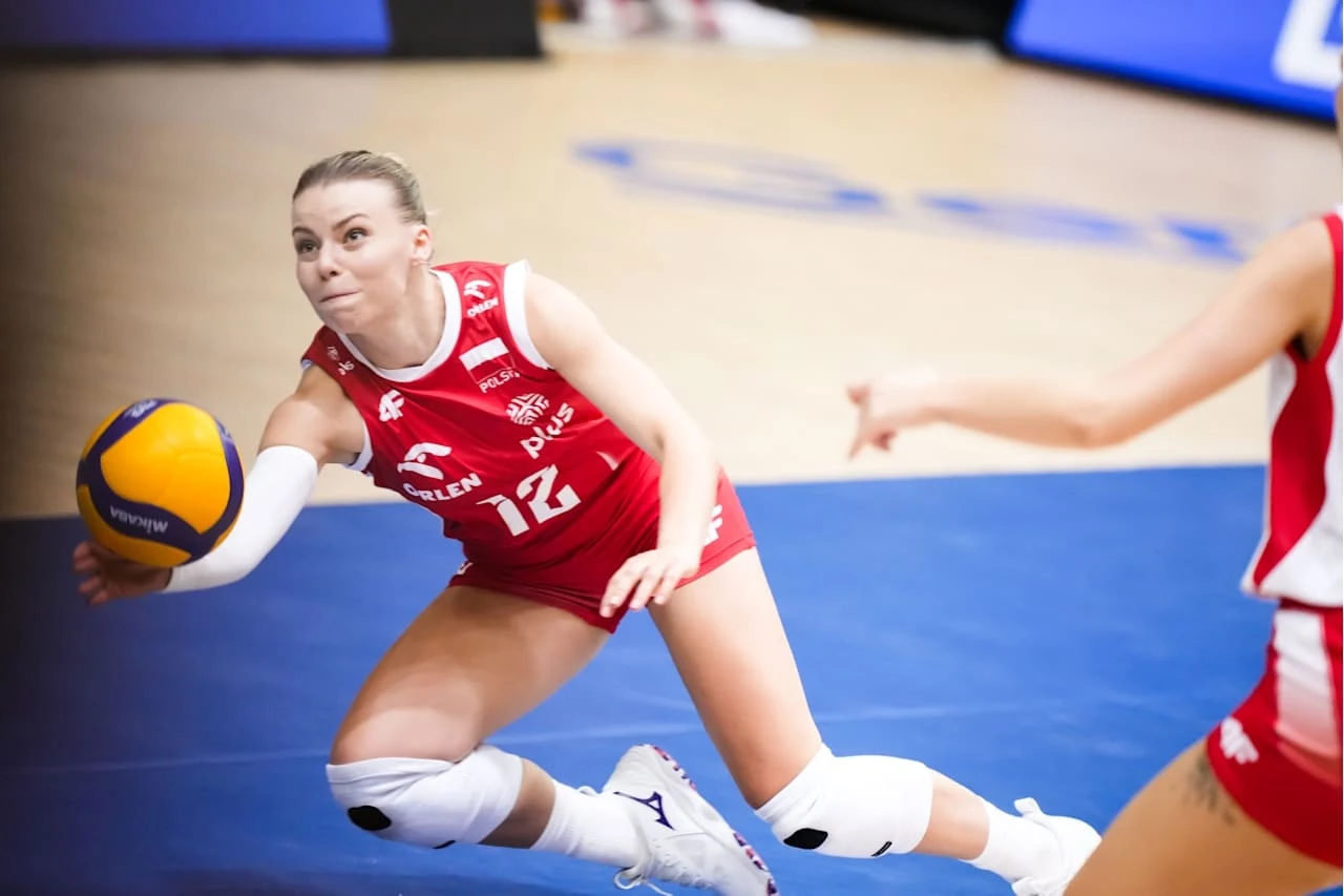 Liga Narodów siatkarek. Aleksandra Szczygłowska to podstawowa libero reprezentacji Polski Siatkarka w czerwonym stroju z numerem 12 wykonuje dynamiczny ruch podczas próby przyjęcia piłki, skupiona na grze, w tle fragment innej zawodniczki i boisko sportowe.