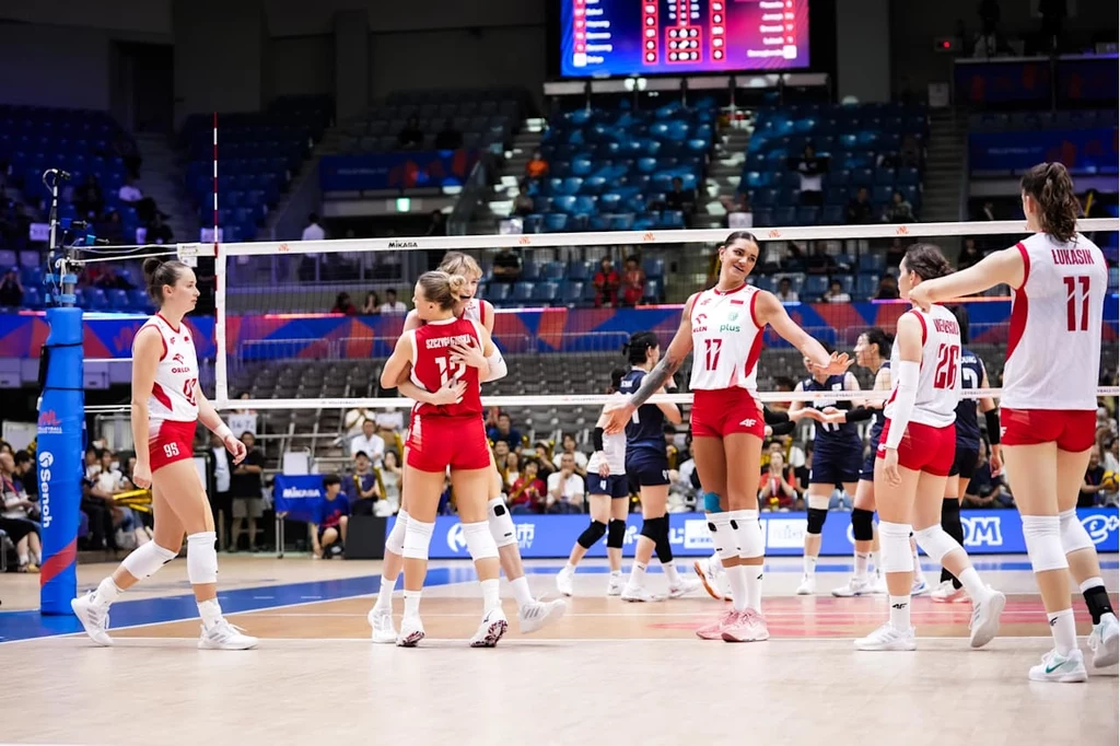 Grupa siatkarek w biało-czerwonych strojach świętuje punkt zdobyty podczas meczu na hali sportowej, w tle trybuny z kibicami oraz tablica wyników.