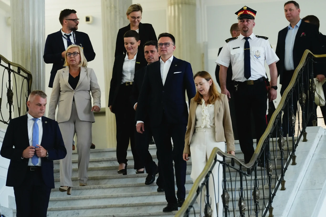 Grupa osób w formalnych strojach schodzi marmurowymi schodami w przestronnym, jasnym wnętrzu budynku rządowego, wśród nich widać także funkcjonariusza w mundurze oraz kobiety i mężczyzn w garniturach.