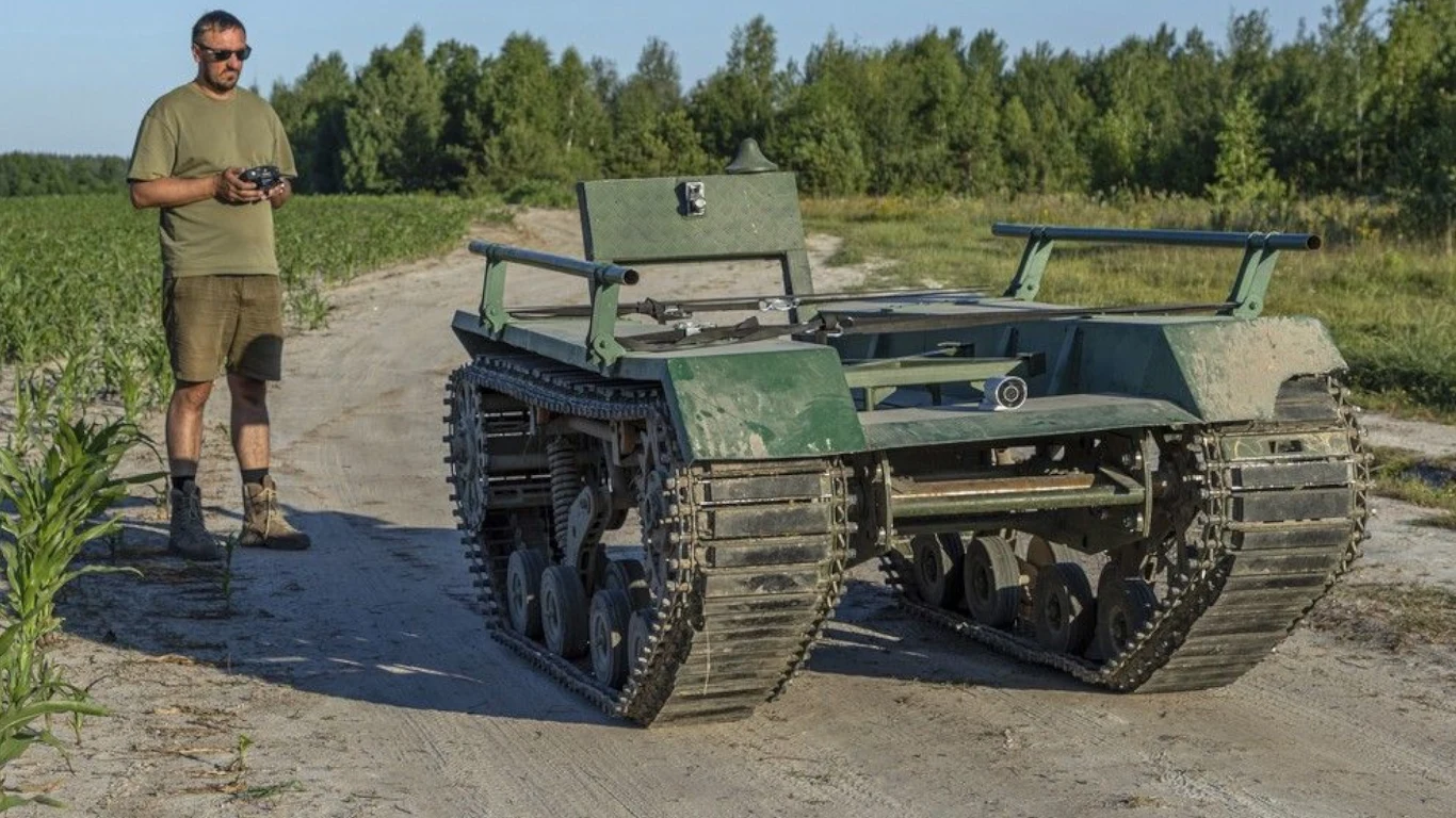 Ukraińcy ranni żołnierze są ewakuowani specjalnymi robotami z pola walki (zdjęcie ilustracyjne) Mężczyzna w krótkich spodenkach i okularach przeciwsłonecznych sterujący zdalnie robotem gąsienicowym na polnej drodze, w tle pole oraz drzewa.