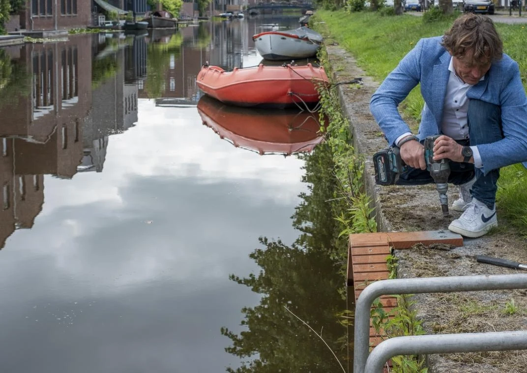 Specjalne schodki zainstalowano wcześniej już w miejscowości Amersfoort (prowincja Utrecht) Mężczyzna w niebieskiej marynarce używa wiertarki przy brzegu kanału, obok pomarańczowa łódka przycumowana do lądu, w tle spokojna woda z odbiciem pobliskich budynków.