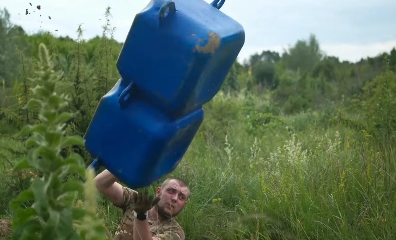 Nowe pontony z plastiku. Ukraińcy budują z nich przeprawy w kilkanaście minut Mężczyzna w mundurze wojskowym pokonuje przeszkodę z dużym, niebieskim pojemnikiem podczas ćwiczeń w zarośniętym terenie