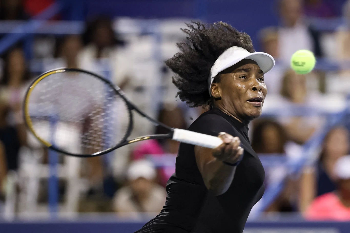 Venus Williams Tenisistka w czarnym stroju wykonująca dynamiczne uderzenie, trzymająca rakietę z pełnym zaangażowaniem, piłka tenisowa znajduje się w powietrzu, tło rozmyte z widownią stadionową.