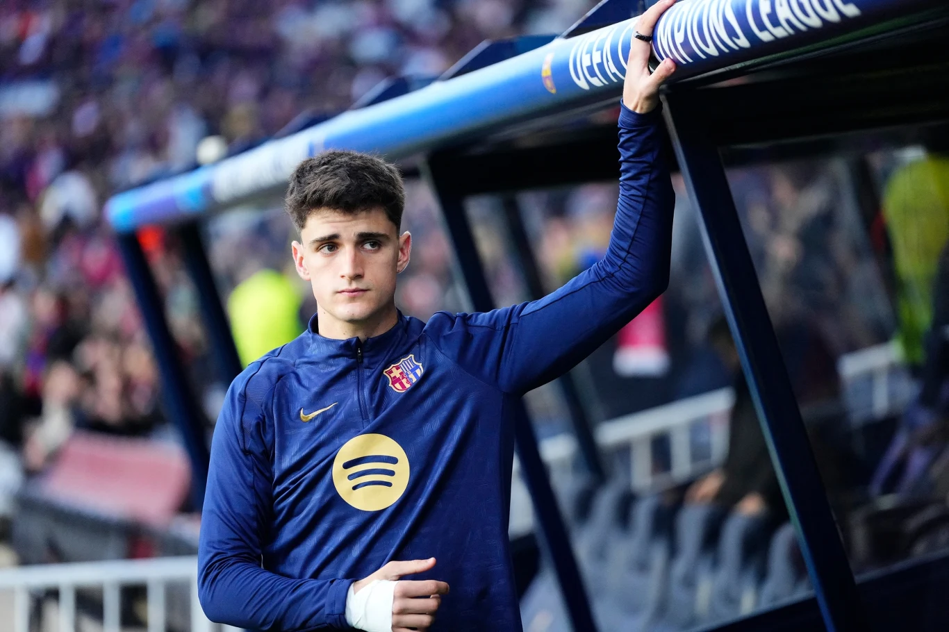 Młody piłkarz w granatowej koszulce klubowej z logo sponsora stoi przy ławce rezerwowych, trzymając rękę opatrzoną bandażem na oprawie ławki, w tle stadion z kibicami.