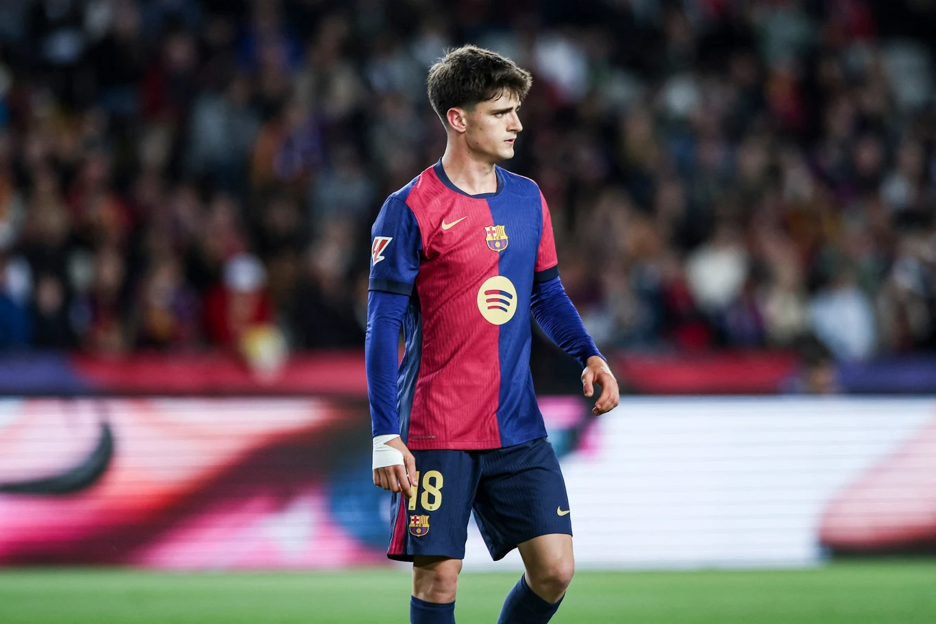Młody piłkarz w barwach FC Barcelona stoi na murawie stadionu z bandami reklamowymi w tle, skupiony, wokół rozmyta publiczność.